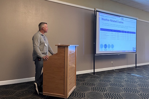 A man speaks standing behind a podium, slide with 'Weather Related Crashes' displayed on a projector screen on the right.